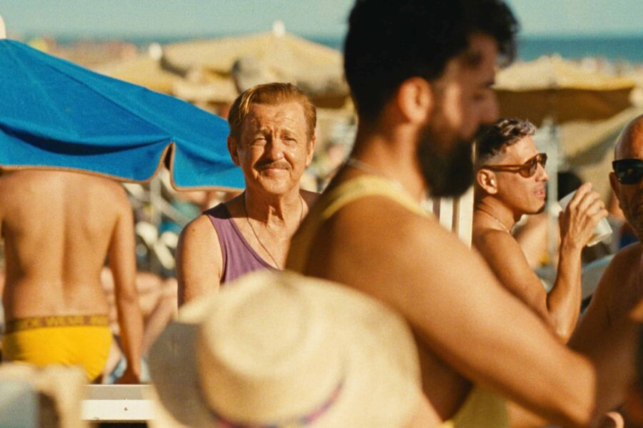 A crowded beach with people facing to the side and away from the camera, one person is in focus and looking down the lens of the camera.