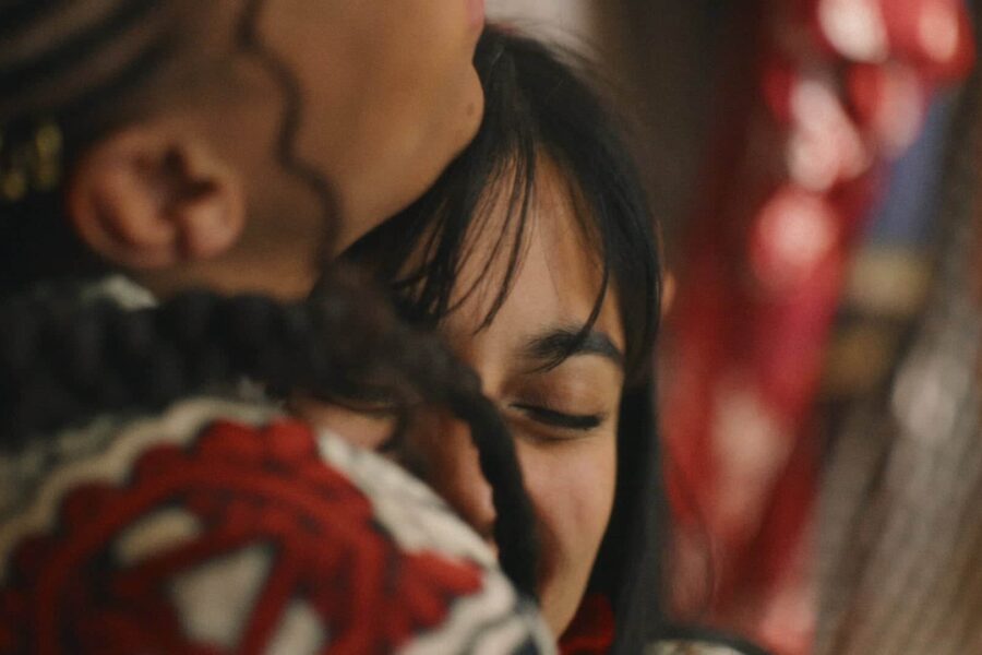 A close up of two people embraced, you can only see the shoulder and chin of the person on the left as the shot is tight, the person on the right has dark hair, and is nuzzled into the hug, smiling.