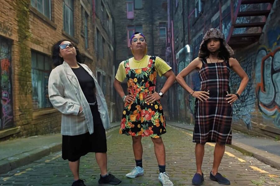 An image with three people stood facing the camera, the person on the left is wearing a black skirt and a long grey coat, the person in the centre is wearing a bright yellow t shirt, a floral pair of dungarees. The person on the far right is wearing a tartan patterned dress, trainers, and a fluffy hat.