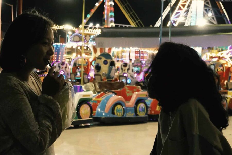 An image with two people looking at each other, they are primarily in dark, behind them is a bright carnival with a carousel and bumper cars in bright colours.