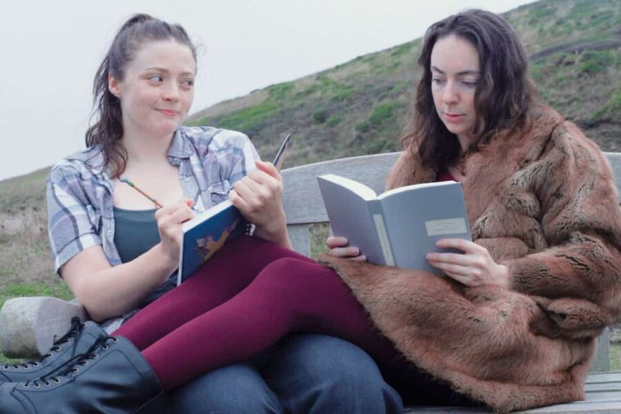 Two people sat on a bench in front of a hill- the person on the left has a pony tail and is holding a book, the person on the right is in a large fur coat and red rights, they are also reading a book.