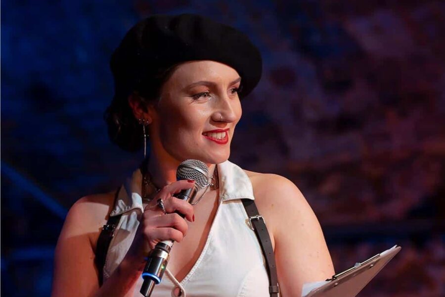 A person stood holding a microphone and holding a clipboard. They are facing out to an audience who are not pictued.