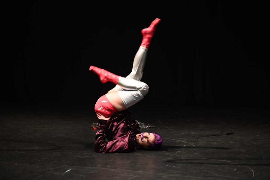 Aperol lays on their back on a stage. They have purple hair, a wonderful maching purple handlebar moustache. He's wearing thigh hi silver leggings, pink socks with little white lightning bolts and matching underwear