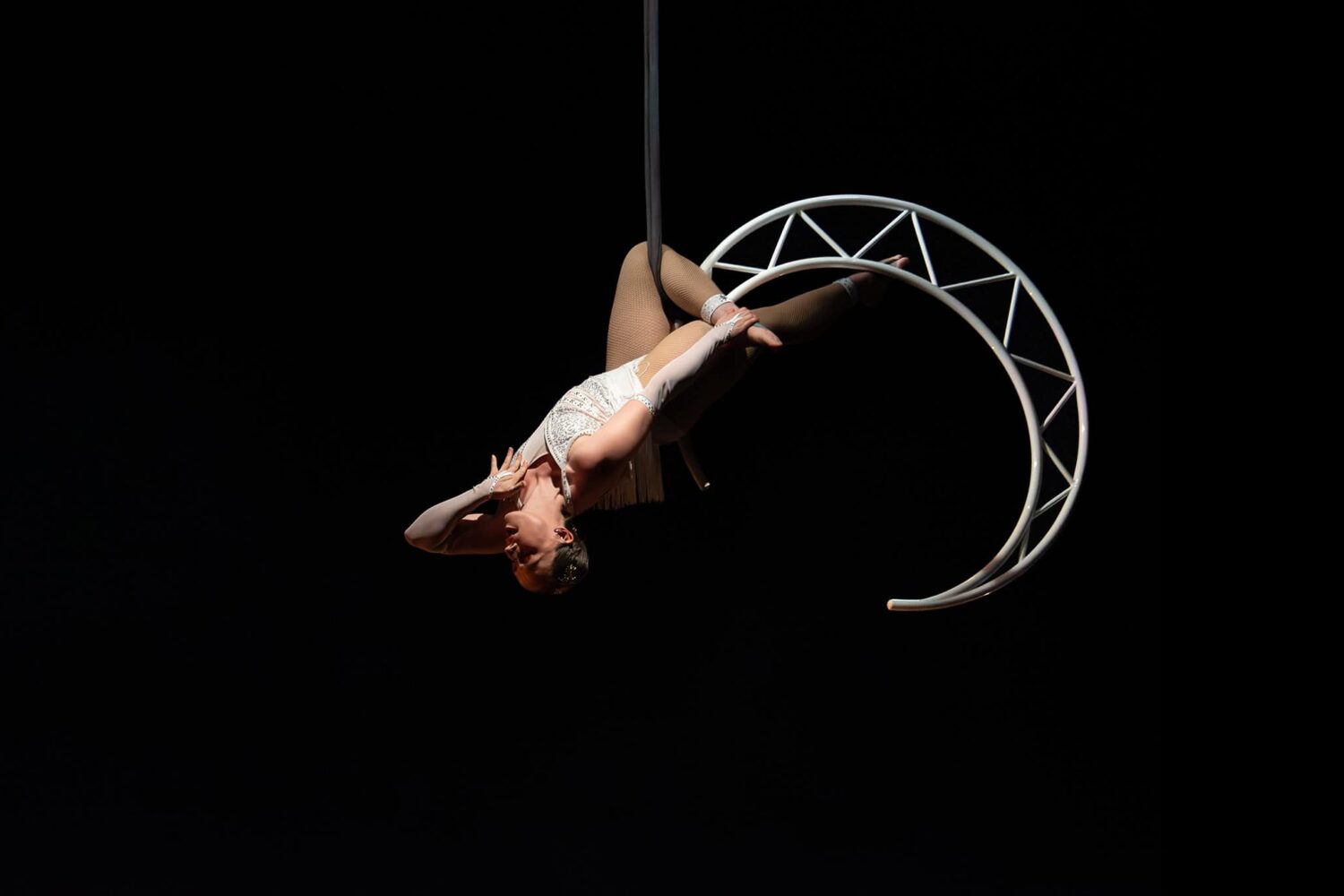 An aerial artists soars in the air extending their body from a crescent moon shaped trapeze