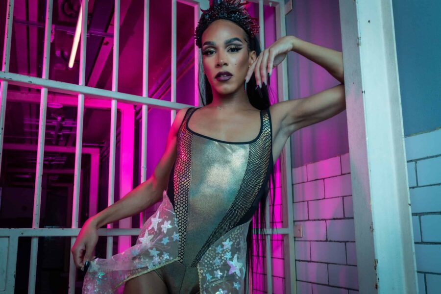 Posed leant against a wall with jail cell bars behind them, Dominique is wearing a silver and black leotard outfit.