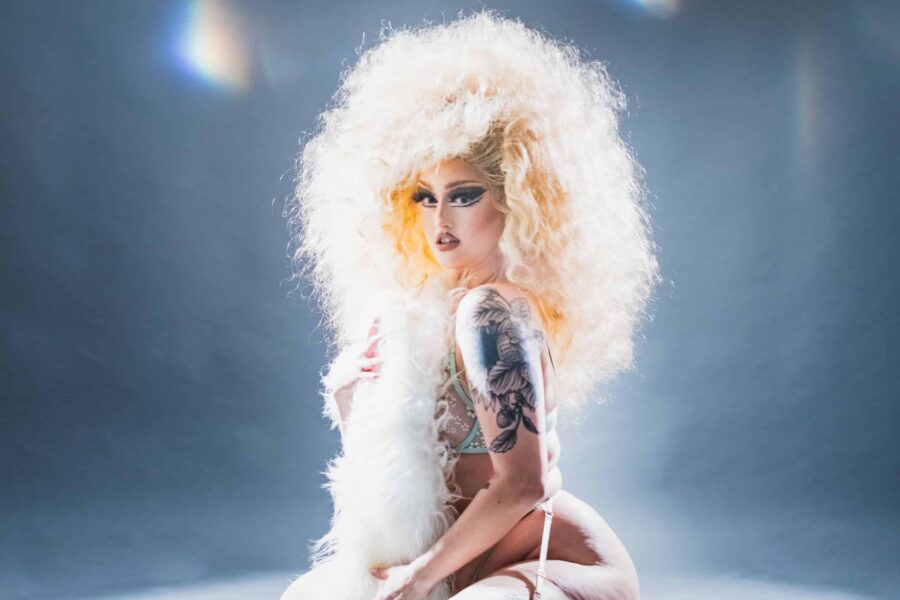 Kitten Fleek sits on her knees, her body sideways ber her head facing the camera and her expression innocent. Her blonde wig is huge and puffs around her like a pool of light.