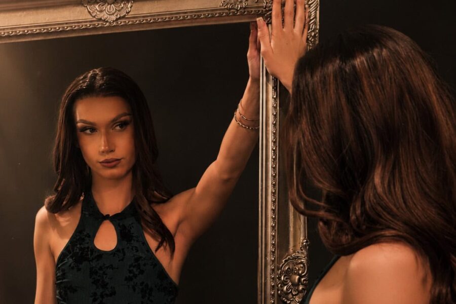 Lipsy is captured admiring themself in the mirror, leant against it, wearing a black mini dress and gold jewellery.