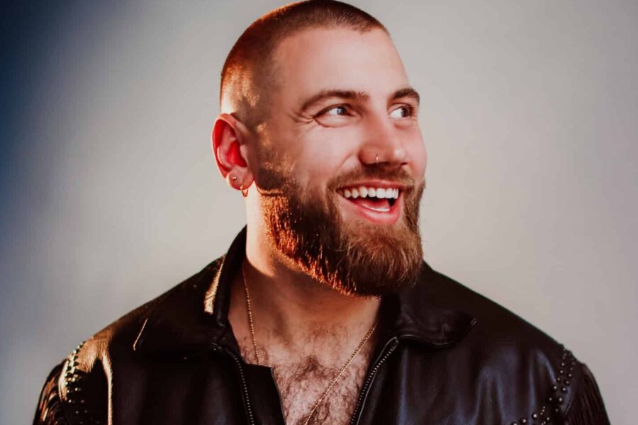 A photo of Morgan Rees standing infront of a grey background. Morgan is wearing a leather jacket, seems to have forgotten his shirt so is showing off his chesthair! He's looking to the left and smiling with a beard and very short cropped hair.
