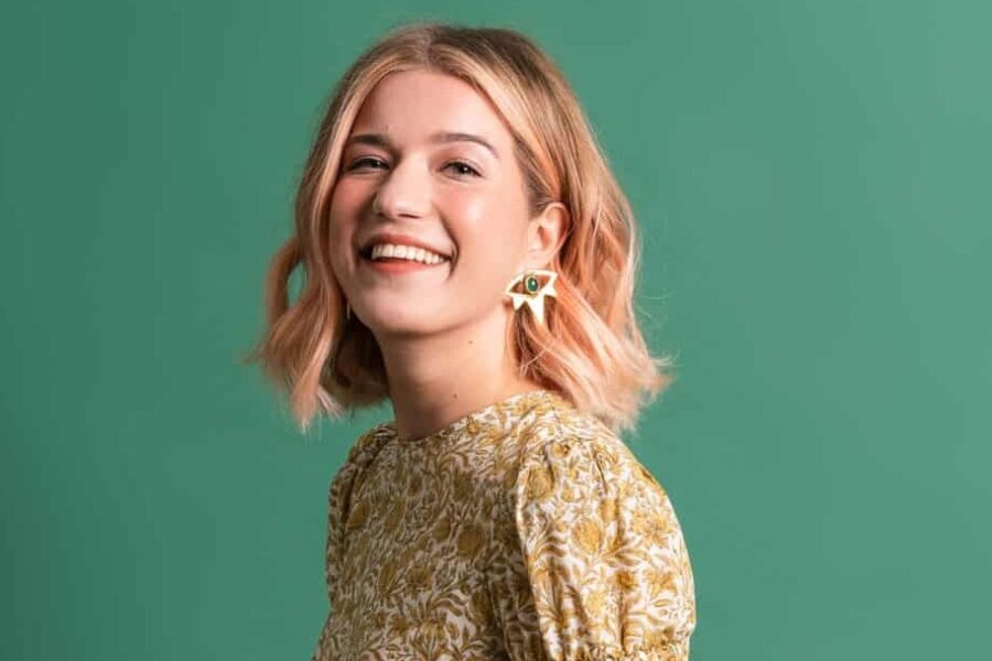 Olga sits sideways on a wooden stool looking a the camera. She is wearing a flower print dress and holds a bunch of sunflowers on her lap.