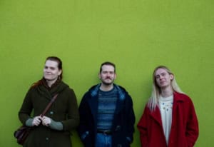 an image of bearpit. 3 people stand in front of a green wall background