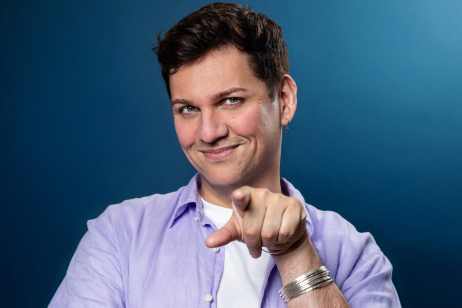 Dane stands infront of a blue background wearing a lilac overshirt and white Tshirt. Arms folded he is pointing down the camera lens and smiling