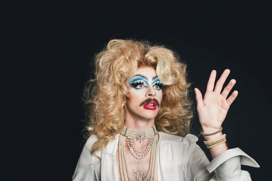 YuGiHoe is looking dreamily at their own hand in a flowy Stevie Nicks Esque white robe, their hair is blonde and flowing and their blue eyeshadow is bright and bold.