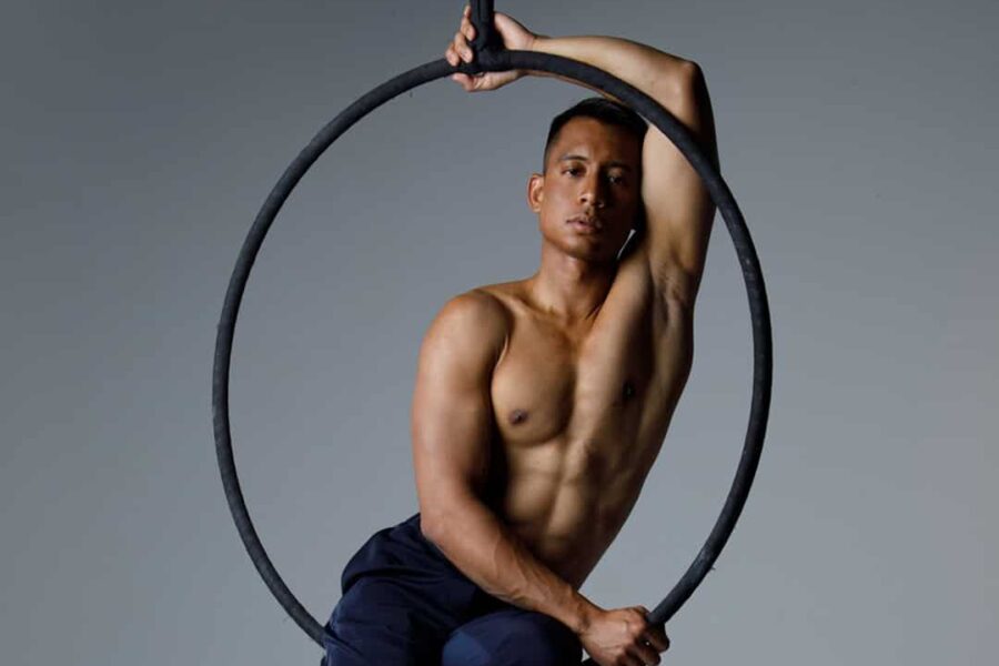 An aerial performer sat in a hoop in front of a grey background, their full body is visible.
