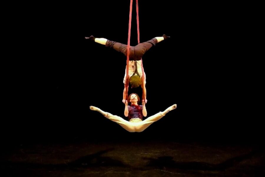 And image on a black background. A person is upside down suspended by red ropes, legs aside. They are holding another person by the arms who is lifting themselves up