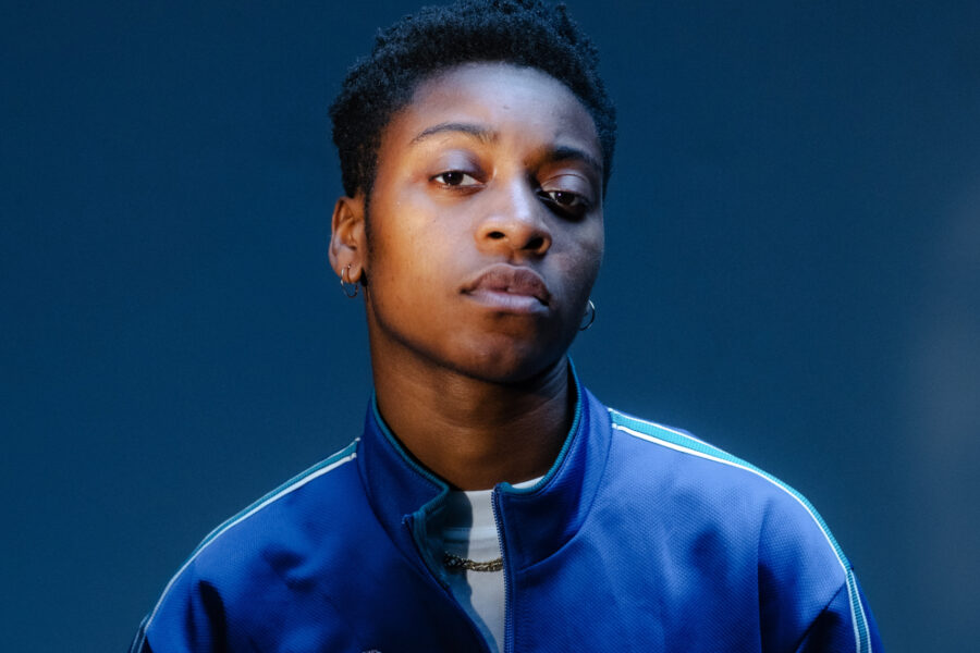 Nat Oakes stands under moody lighting on a dark blue background. They are wearing a blue sports jacket, white tee and gold chain.