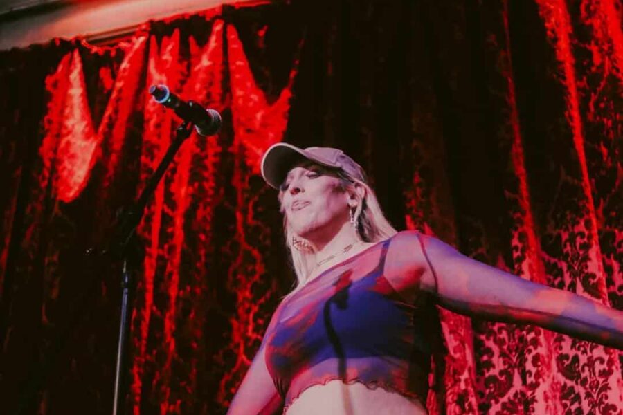A person with long blonde hair and a hat is stood in front of a red curtained background. There is a microphone on stage they are singing into.