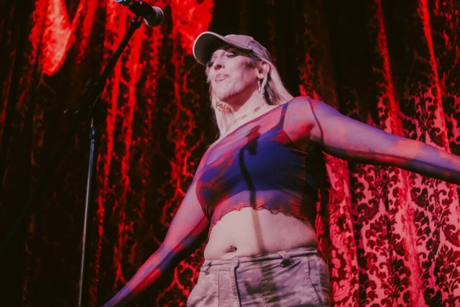 A person with long blonde hair and a hat is stood in front of a red curtained background. There is a microphone on stage they are singing into.