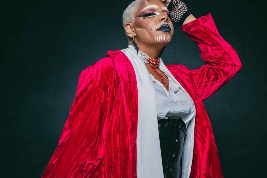 A person in front of a black background wearing a bold and textured red suit, open at the front. Striking a bold pose.