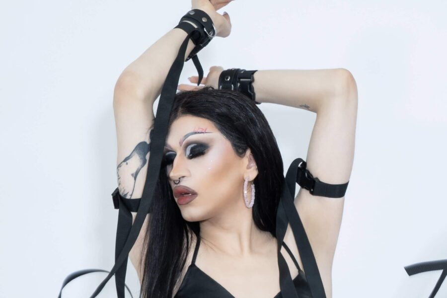 A person with long black hair and big makeup is centre on a white background, looking off to the side. There are ribbons around the person's wrists and arms that flow off out of shot.