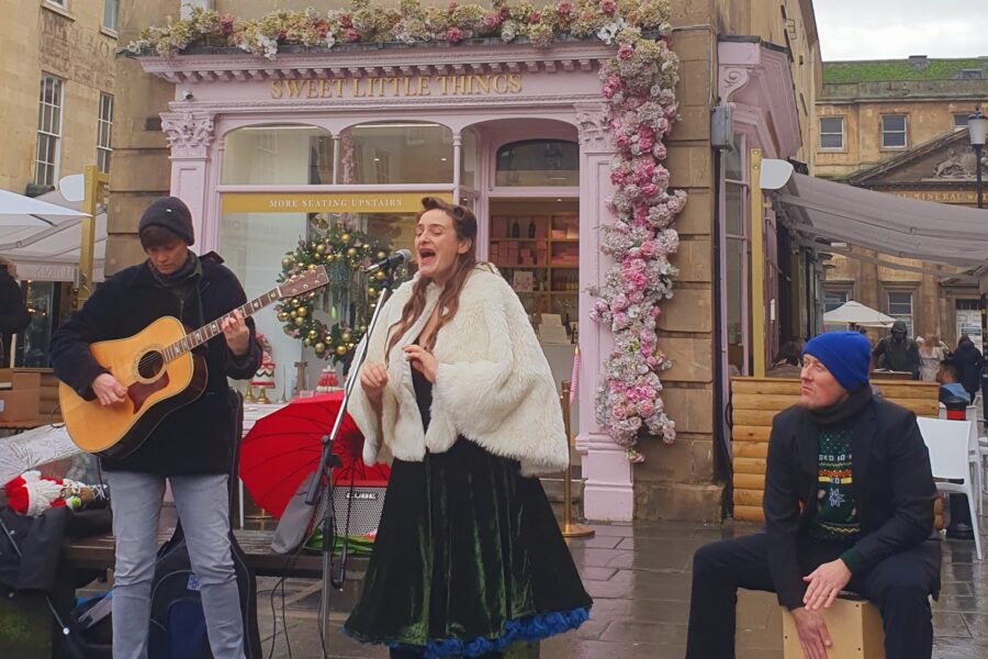 Three people are performing on the high street, on the far left is a person in a hat playing the guitar, in the middle is a person in a green dress and red boots singing into a microphone, on the right a person in a beanie playing a cajon drum.
