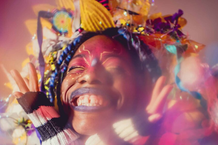 A mid-shot of a person with their head in their hands, they are wearing striped fingerless gloves. They are wearing an etravagant textured headdress with yellow flower petals and blue ribbons. The person is looking up, with their eyes shut, and there is pink and goldmakeup around their eyes.