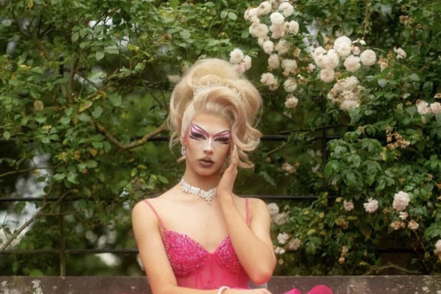 A person sat on a bench infront of a hedge of leaves and white flowers, the person is dressed in a big pink gown. Their hair is in a glamorous blonde updo and their head is rested in one of their hands.