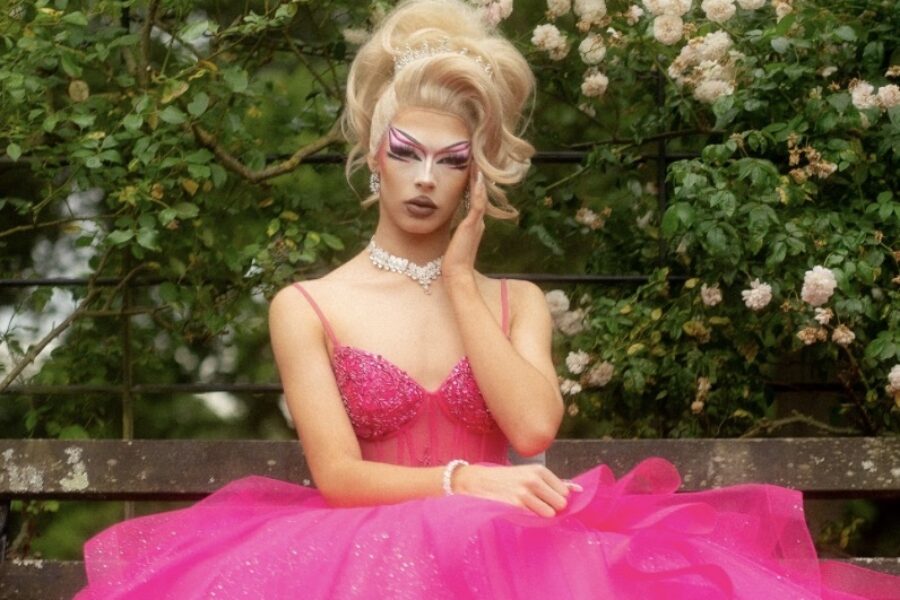 A person sat on a bench infront of a hedge of leaves and white flowers, the person is dressed in a big pink gown. Their hair is in a glamorous blonde updo and their head is rested in one of their hands.