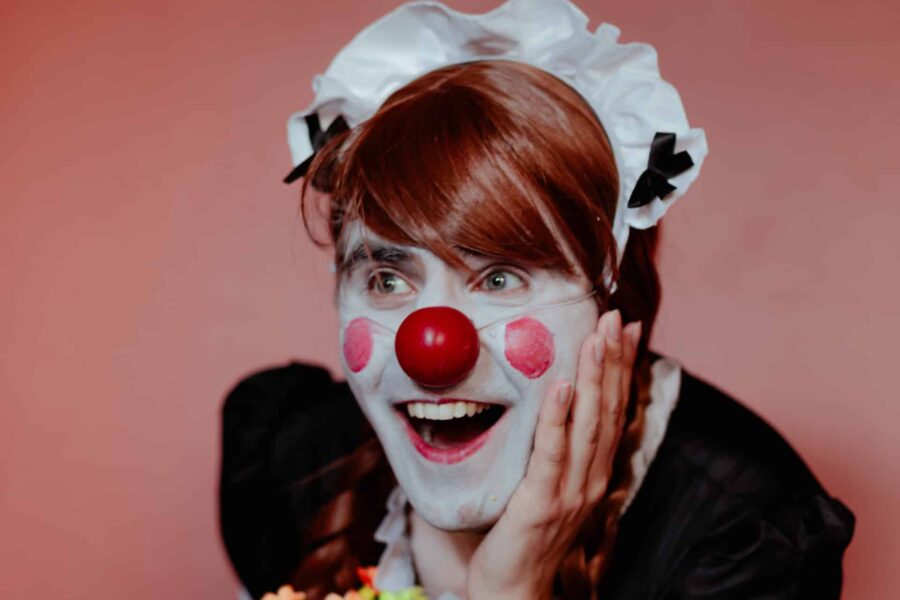 A person in front of a pink background wearing a maid outfit. They are leant forward, holding a bunch of flowers in one hand and clutching their face in the other. They have on white face make up, pink circles on their cheeks, and a red nose. They are smiling at something beyond the camera.