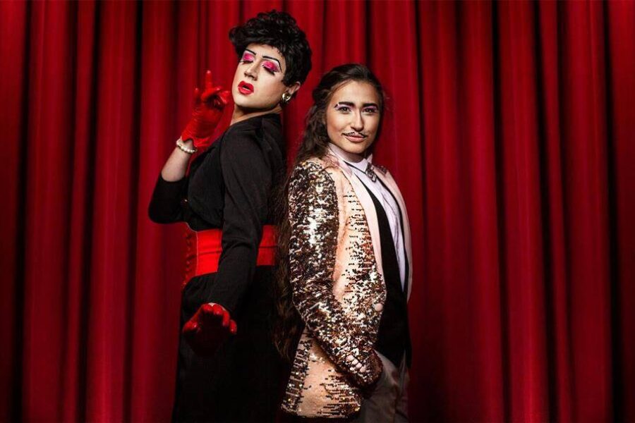 A drag queen in a black dress and large red belt stands with a Drag King in a gold sequin jacket and pencil moustache in front of a red velvet curtain