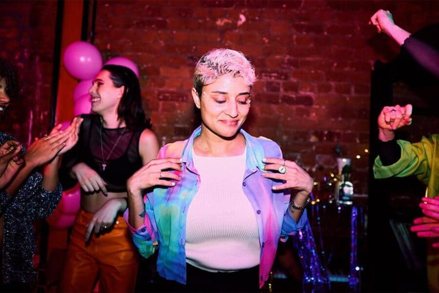 It looks like a fun party is happening, people laugh in the background, there are pink ballons. There is a person centred in the image wearing a blue jacket and white top with short bleached hair with contrasting roots.