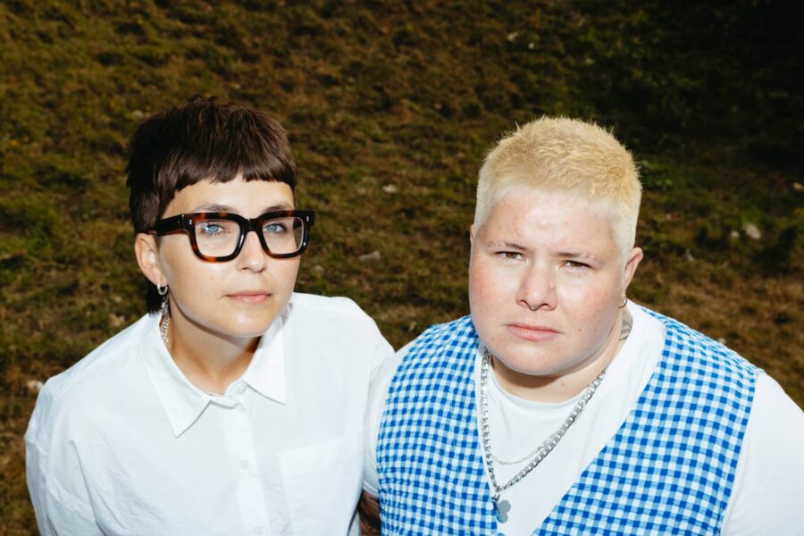 Image of ARXX band with Hanni on the left in a white shirt and thick black glasses and short brown hair. Clara is on the right with a blue and white check top and short bleach blond hair.