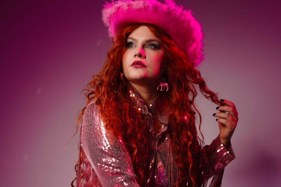 Laura Batten crouches on the floor surrounded by disco balls and dressed as Chappell Roan with curly red hair and pink fluffy cowboy hat