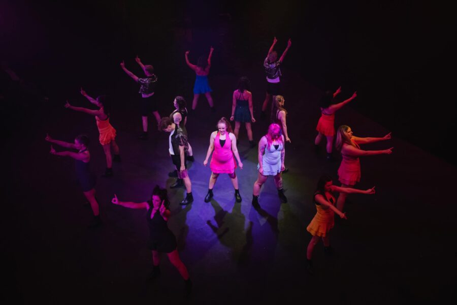 A group of people performing on stage, the lights are blue tinted and dim. They are in two circles, one surrounding the other. Boldly posed with hands balled by their sides.