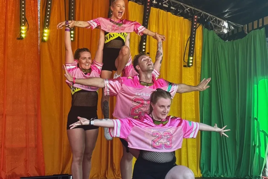 A group mid-performance in front of a rainbow backdrop. They are in a totem style position, lined in varying levels of height, there is a person at the back who is being lifted by two others. They are wearing bright pink tops.