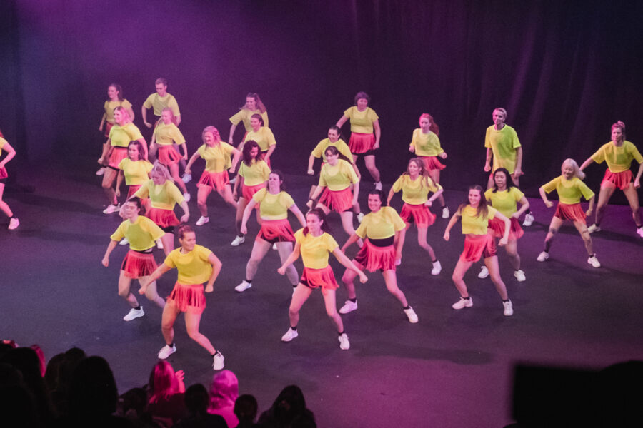 A big performance group on stage, organzied into a triangle, with purple stage lights shining onto them. They are wearing matching pink shorts and bright yellow t-shirts.