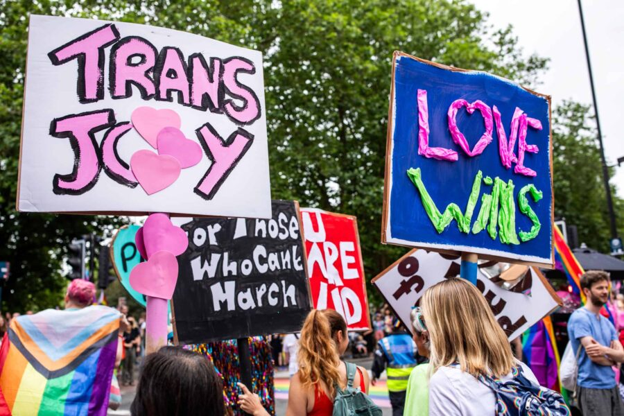 An image of people holding up signs and placards for pride. They are in front of trees, the visible placards read 