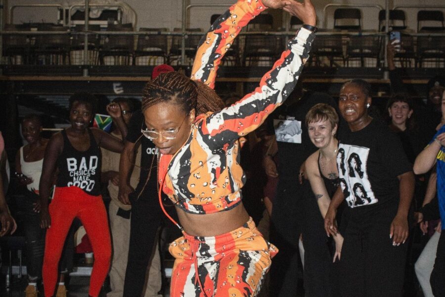 A person mid-dance. They are doing an impressive dance move, with both their arms extended behind them. They are wearing an orange tracksuit style outfit.