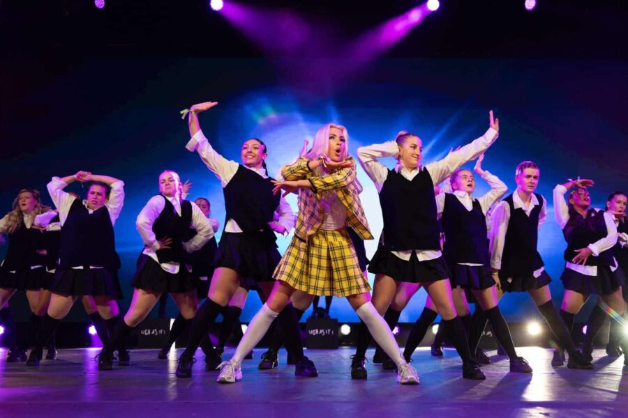 A photo taken mid-performance. In the centre of the photo is a young performer with long blonde hair and a yellow plaid outfit. Around them is a group in matching black and white outfits.