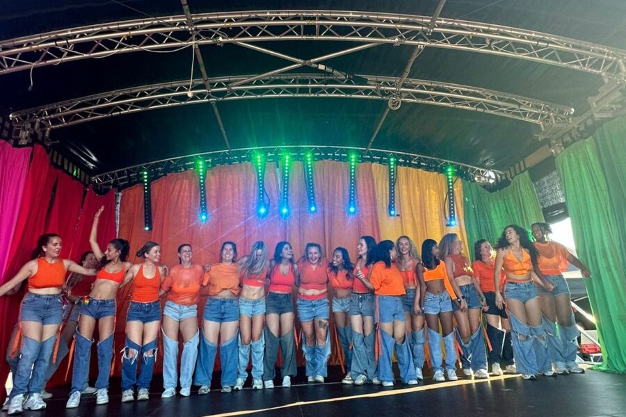 A group mid-bow on the pride dance stage, in front of a rainbow back drop. They are wearing matching orange tops and blue jeans.