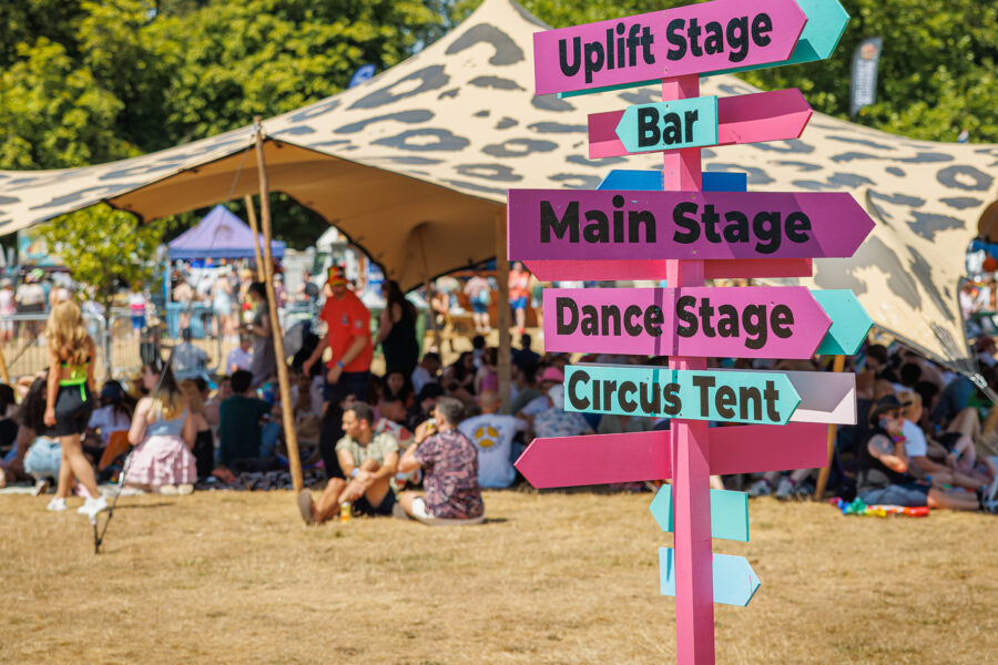 A way finding sign with multiple direction signs on it stating Dance stage, main Stage, bar, Circus Tent. The background a leopard spot stretch tent