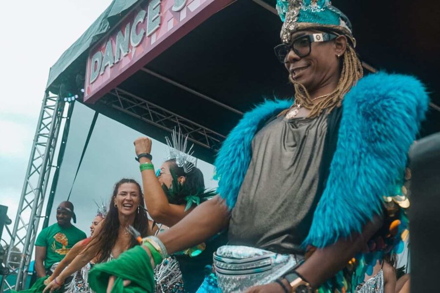 A photo mid-performance, the focus of the picture is a dancer in an elaborate carnival costume, with blue feather shoulder accents and a blue feather head piece. Others dance on stage behind them.
