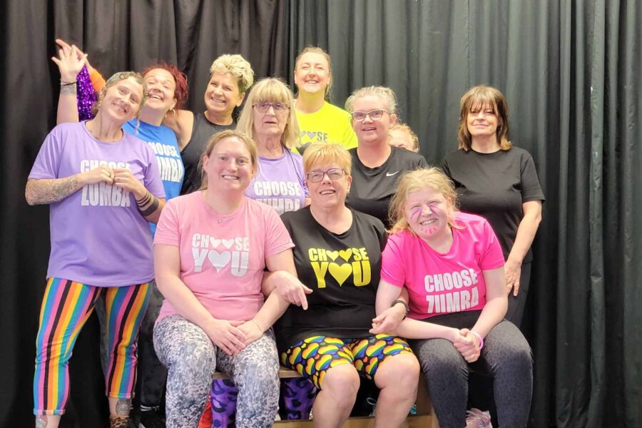 A group lined up for a photo in front of a black curtain backdrop. They are wearing pink, yellow, black, and purple shirts that read 