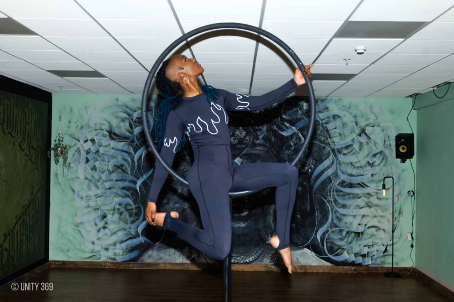 A performer posed in an aerial hoop, they are in an all black gymnast outfit, and their head is tilted up to the ceiling.