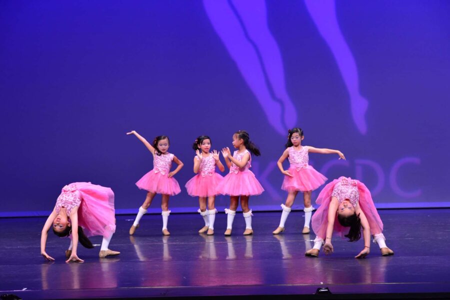 A group of young dancers infront of a blue backdrop on stage. They are mid performance, all wearing pink tu-tu like costumes with white socks. Two of them are in back-bends and the rest are posed elegantly with their arms out.