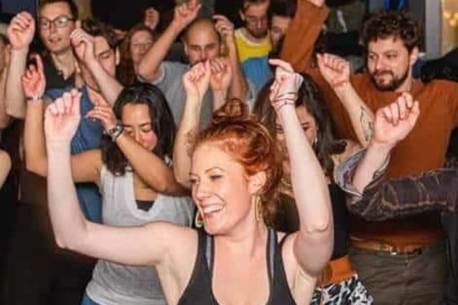 A group of people mid-dance, they have their hands in the air, mid-click. They are all smiling.