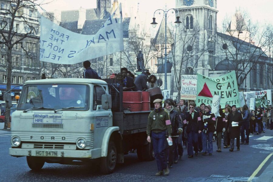 An archive image of marchers supporting the Gay News appeal against its conviction in 1978-79 a flat bed truck has people playing steel drums on it whilst people march alongside with pink armbands and banners that read National Gay News Defence Committee