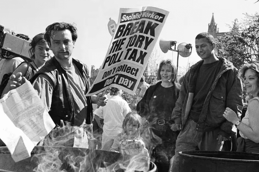 A black and white image of people stood in protest by barrels. The barrels are smoking and papers are being burnt in them. A person with short hair is stood in the foreground holding a placard that 