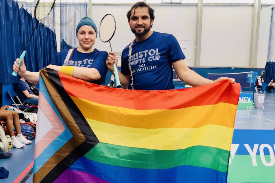 Two people holding a pride flag out, in their other hands are badminton rackets.
