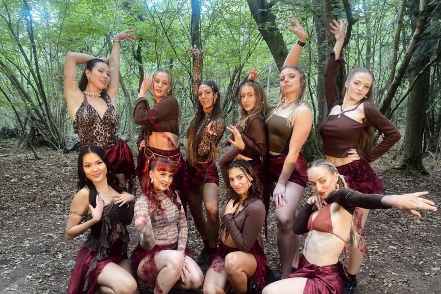A group posed within the woods - they are wearing deep red and leopard print outfits. Posed in two lines and smiling happily at the camera.