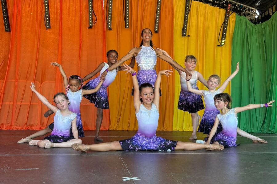A group of dancers, organised into a pyramid shape, posed in front of a rainbow backdrop on stage. They are wearing white and purple dance costumes, and have their arms stretched out elegantly.